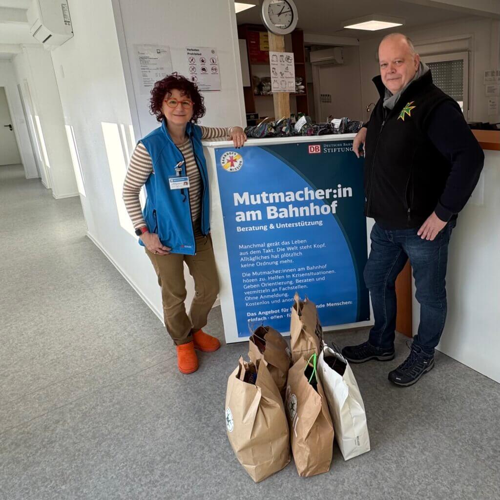 Aufnahme in der Bahnhofsmission Frankfurt am Main. Links Anja Wienand (Leiterin der Bahnhofsmission) und rechts Dirk Augustin (St. Lazarus-Orden) mit gespendeten handgestrickten Socken vor einem Werbeaufsteller der Bahnhofsmission.